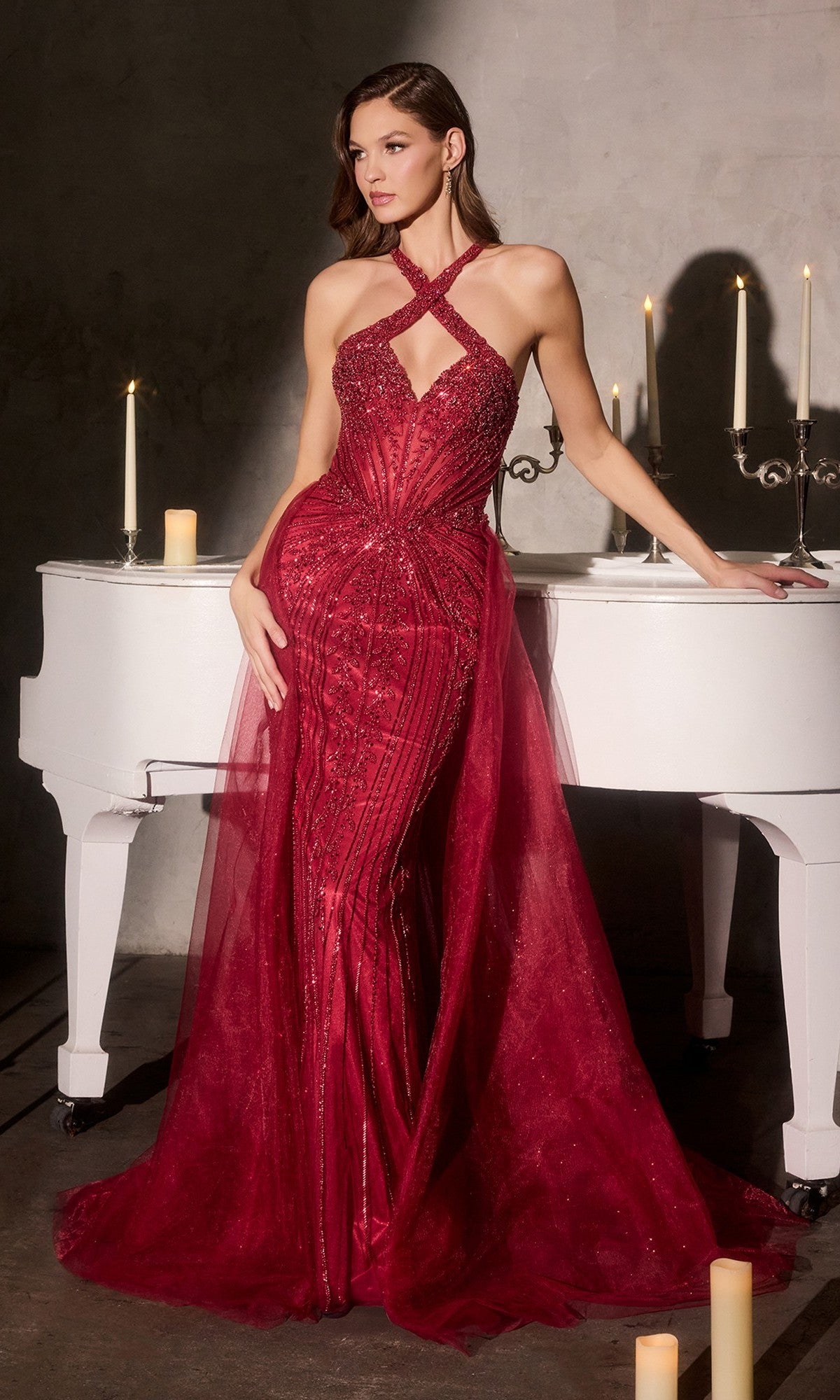 A woman wears the Ladivine CC495 Long Prom Dress in deep red, featuring a fitted beaded design and crisscross halter neckline, standing by a white grand piano with softly glowing candles in the background.